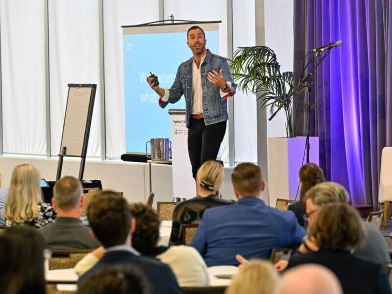 Photo taken during the Together25Ensemble conference featuring a man in a suit speaking on stage