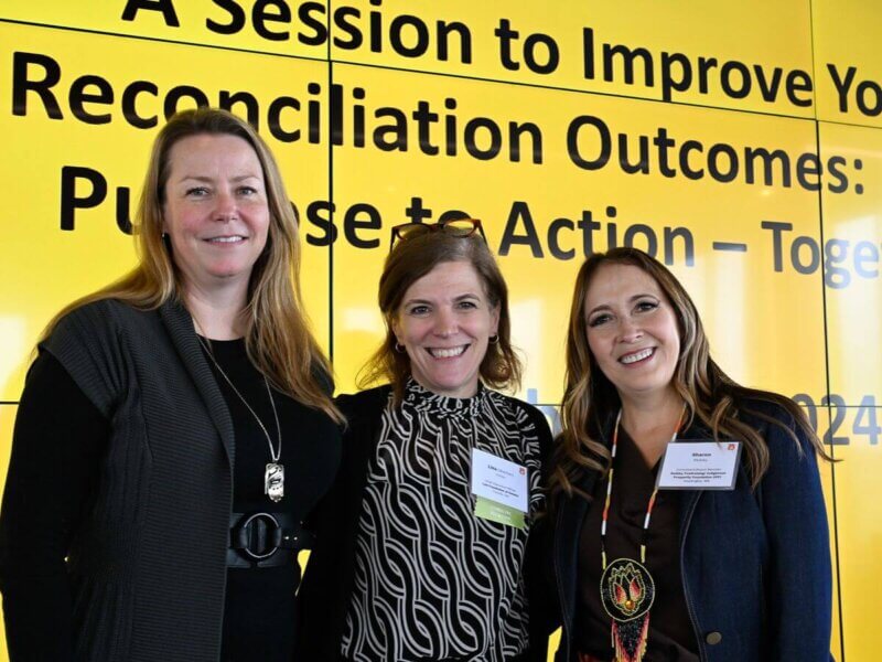 Photo taken during the Together25Ensemble conference featuring three women posing in front of a yellow background
